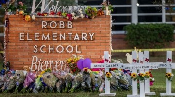 The welcome sign at the front of a school that says "Robb Elementary School." The sign is surrounded by a memorial of flowers and crosses with children's names.