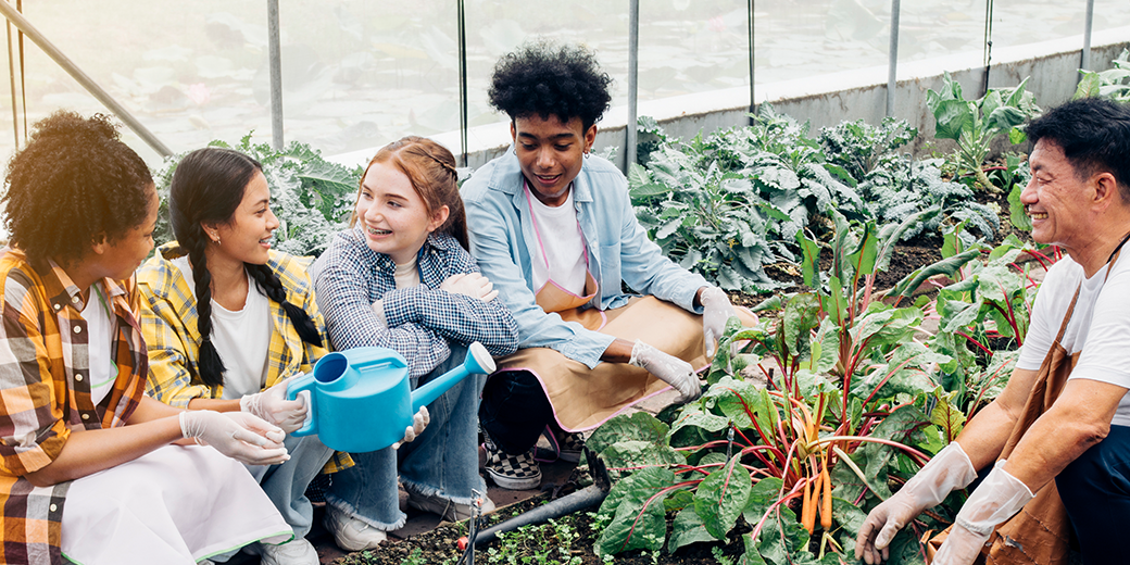 4 smiling high school students crouch down next to a garden row filled with plants. One holds a watering can. Across the row, an older teacher wearing gloves and an apron smiles at the students.