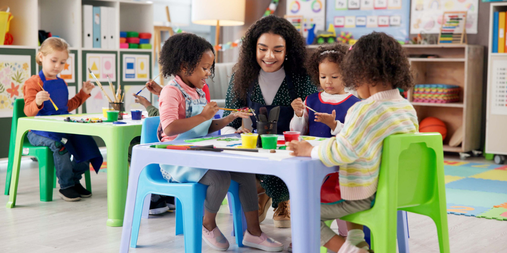 A teacher smiles while guiding four young children in a painting activity at colorful tables in a preschool classroom, with art supplies visible in the background.