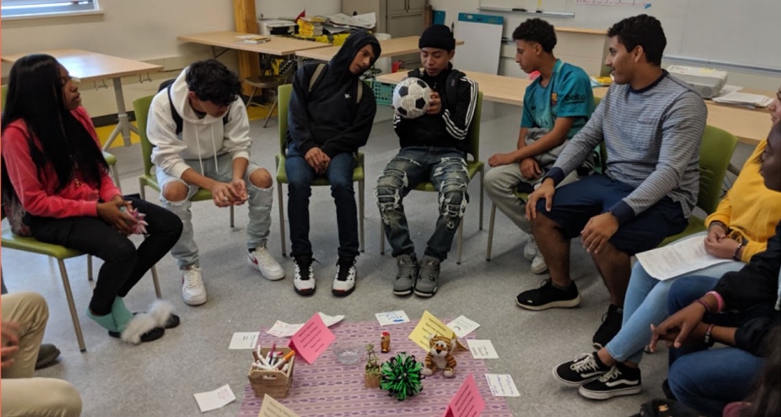 Students participate in a restorative circle, sitting in chairs in a circle. One student holds a soccer ball and the other students are paying attention to him.