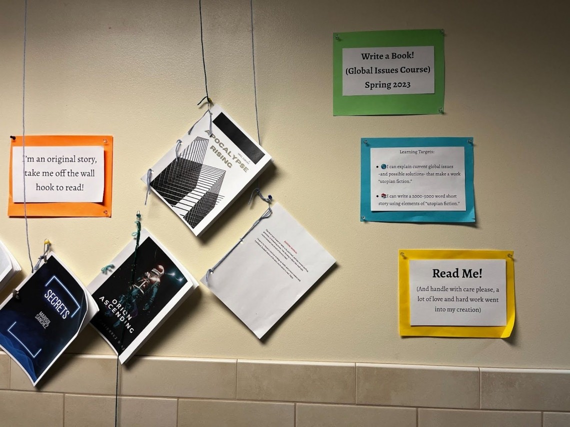 A photograph of the wall in a school hallway where passerby can read student work. Posters explain the work is from the “Write a Book! (Global Issues Course) Spring 2023,” share the learning targets of the class, and tell people to “Read me!” Next to the posters hang examples of students’ original stories, booklets of paper with creative covers and titles like “Apocalypse Rising,” “Orion Ascending,” and “Secrets.”