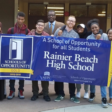 A group of diverse high school students stand in front of a school building, holding up a blue banner with white and yellow lettering: "A schools of opportunity for all students: Rainier Beach High School."