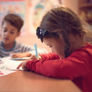 Two young students sit at a classroom table, drawing on paper with markers 