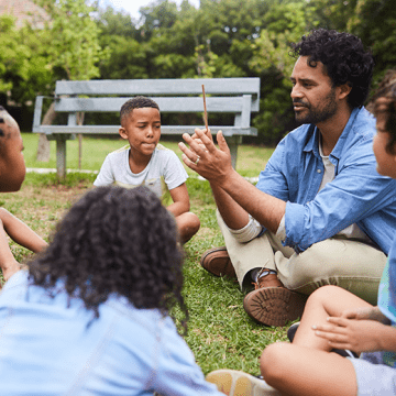 Teacher sitting with young students on lawn by a bench