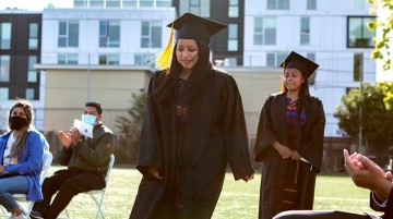 High school students in graduation attire walking outdoors while the audience applauds.