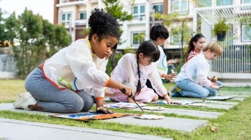 Elementary school students kneel on grass outdoors working on colorful paintings
