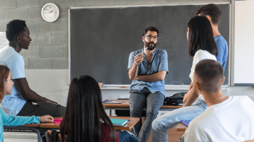 A teacher at the front of the classroom with high school-aged students listening to him speak.