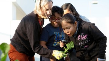 A teacher holding a plant while students view it with a microscope.