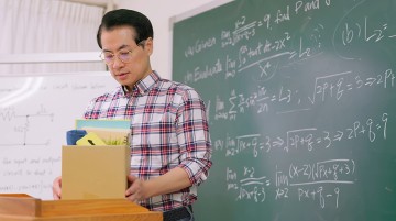 A teacher holds a cardboard box on a desk in front of a chalkboard.