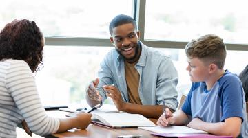 Two adults assist a student with an assignment in a classroom.