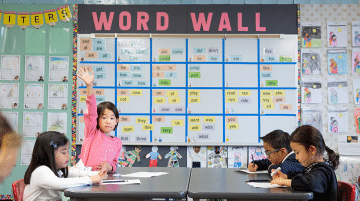 An elementary student raises their hand at a shared classroom table.