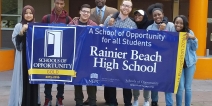 A group of diverse high school students stand in front of a school building, holding up a blue banner with white and yellow lettering: "A schools of opportunity for all students: Rainier Beach High School."