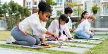 Elementary school students kneel on grass outdoors working on colorful paintings