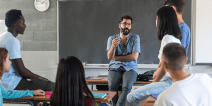 A teacher at the front of the classroom with high school-aged students listening to him speak.
