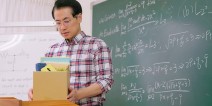 A teacher holds a cardboard box on a desk in front of a chalkboard.