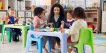A teacher smiles while guiding four young children in a painting activity at colorful tables in a preschool classroom, with art supplies visible in the background.