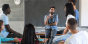 A teacher at the front of the classroom with high school-aged students listening to him speak.