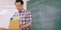 A teacher holds a cardboard box on a desk in front of a chalkboard.